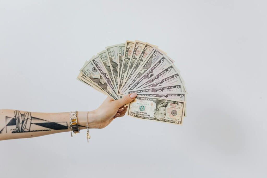 Tattooed arm holding a fan of US dollar bills on a white background, symbolizing wealth and financial success.